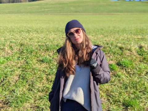 Eine Person mit Sonnenbrille, Beanie und dunkler Jacke steht auf einer Wiese unter einem klaren blauen Himmel. Sie blickt direkt in die Kamera, hat die Hand in der Manteltasche und die Sonne wirft einen Schatten auf den Boden.