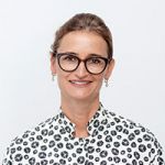Eine Person mit Brille lächelt in die Kamera. Sie trägt ein weißes Hemd mit schwarzem Tupfenmuster. Der Hintergrund ist schlicht weiß.