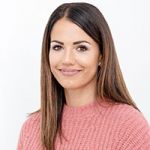Eine Frau mit langen braunen Haaren und einem rosa Pullover lächelt vor einem schlichten weißen Hintergrund.