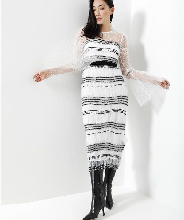 Eine Frau in einem weißen Kleid mit schwarzen Streifen und transparenten gepunkteten Ärmeln posiert vor einer weißen Wand. Sie trägt kniehohe schwarze Stiefel und hat langes, dunkles Haar. Ihr rechter Arm ist erhoben und ruht an der Wand, ihr Gesichtsausdruck ist entspannt.