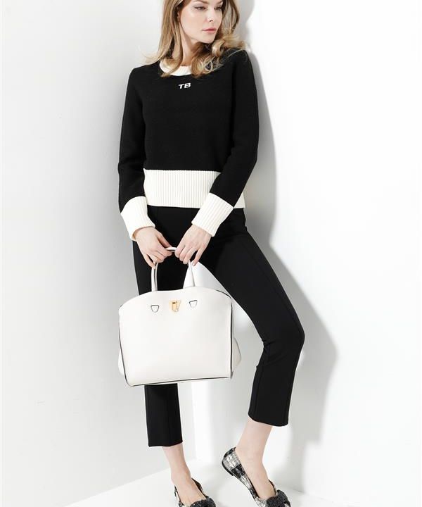 Eine Frau steht vor einer weißen Wand. Sie trägt ein schwarz-weißes Outfit mit Pullover, Hose und Schuhen. Sie hält eine große weiße Handtasche und blickt zur Seite. Ihr Haar ist offen gestylt und das Outfit enthält ein Logo auf dem Pullover.