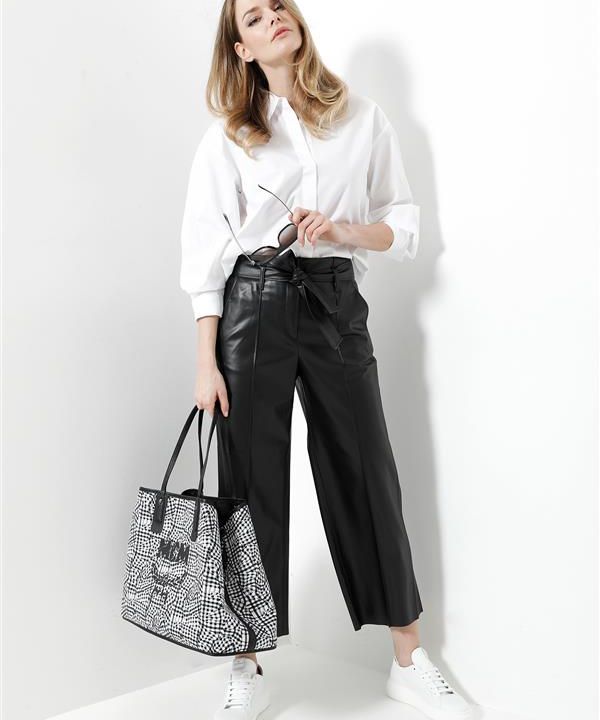 Eine Person in weißer Bluse und schwarzer Hose mit weitem Bein trägt eine Sonnenbrille und eine große gemusterte Einkaufstasche. Sie steht vor einem schlichten weißen Hintergrund und trägt weiße Turnschuhe.