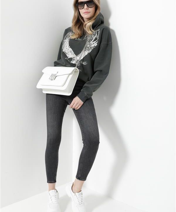 Eine Frau steht vor einer weißen Wand. Sie trägt eine Sonnenbrille, einen dunkelgrünen Kapuzenpullover mit großem Aufdruck, dunkle Röhrenjeans und weiße Turnschuhe. In einer Hand hält sie eine weiße Handtasche.