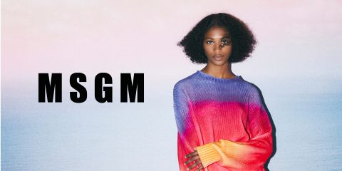 Eine Person mit lockigem Haar steht am Meer und trägt einen farbenfrohen Pullover mit Farbverlauf in Lila-, Rosa- und Rottönen. Die Buchstaben „MSGM“ sind auf der linken Seite des Bildes deutlich zu sehen. Himmel und Meer bilden eine ruhige Kulisse.