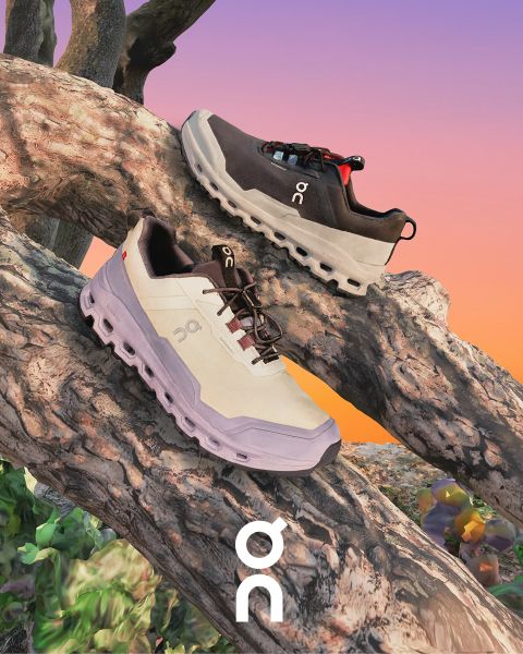 Zwei Sneaker der Marke „On“ werden auf einem strukturierten Ast vor einem farbenfrohen Sonnenuntergangshimmel präsentiert. Ein Schuh ist schwarz mit grauen Akzenten und der andere ist weiß mit dunkleren Details. Darunter ist das „On“-Logo sichtbar.