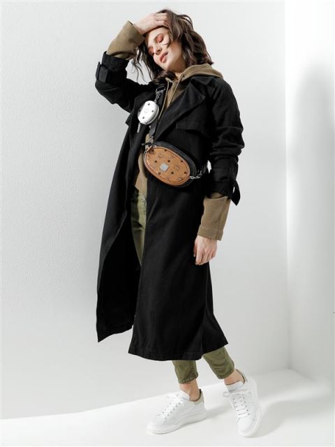 Eine Person posiert in einem stylischen Outfit vor einem weißen Hintergrund. Sie trägt einen langen schwarzen Mantel über einem braunen Kapuzenpullover, olivgrüne Hosen und weiße Turnschuhe. Zu den Accessoires gehören eine Umhängetasche und ein runder Beutel. Die Person stützt eine Hand auf den Kopf.