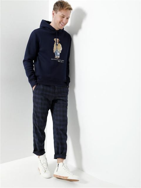 Eine Person steht lächelnd vor einer weißen Wand. Sie trägt einen dunkelblauen Kapuzenpullover mit Bärenmotiv, karierte Hosen und weiße Turnschuhe. Sie hat die Hände in den Taschen und ihr blondes Haar ist lässig gestylt.