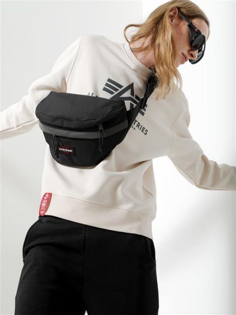 Eine Person mit Sonnenbrille und beigem Sweatshirt posiert mit einer schwarzen Gürteltasche über der Schulter. Sie hat lange Haare und trägt eine schwarze Hose mit einem roten Etikett an der Seite.