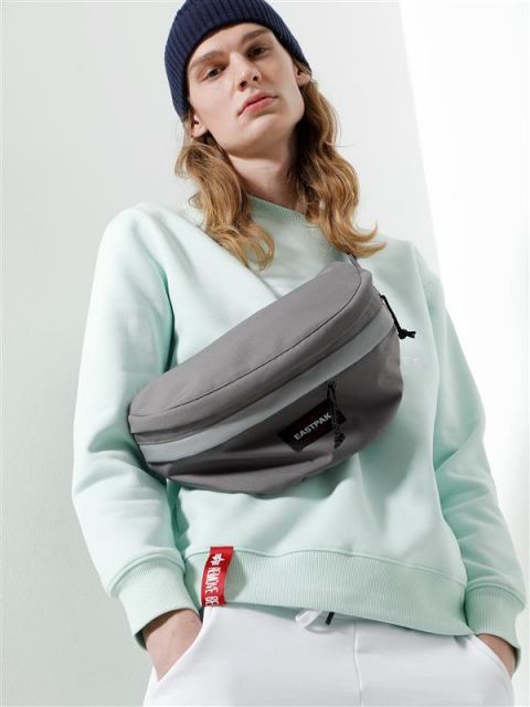 Eine Person mit langen Haaren, die eine blaue Beanie, ein mintgrünes Sweatshirt und weiße Hosen trägt, steht vor einem hellen Hintergrund. Sie trägt eine große graue Gürteltasche quer über der Brust.