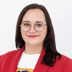Eine lächelnde Person mit langen dunklen Haaren und Brille, die einen roten Blazer über einem weißen Hemd trägt, steht vor einem schlichten weißen Hintergrund.