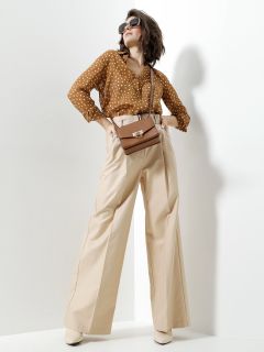 Eine Person posiert selbstbewusst in einer braunen Bluse mit Tupfen, beigen Hosen mit hoher Taille und weißen Schuhen. Sie trägt eine Sonnenbrille und eine braune Umhängetasche und steht vor einem minimalistischen weißen Hintergrund.