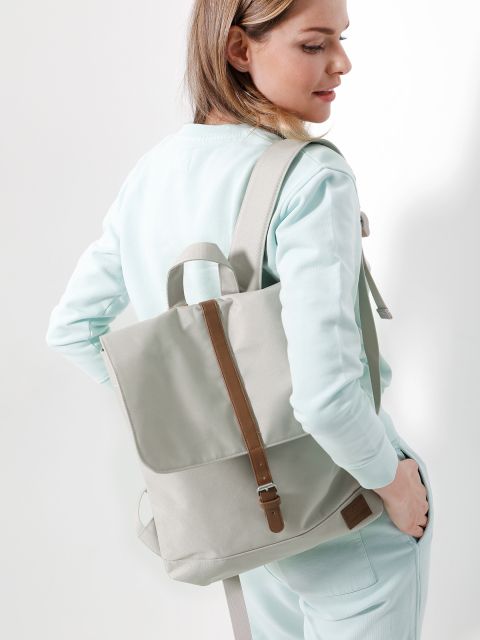 Eine Person in hellblauem Outfit und einem hellgrauen Rucksack mit braunen Riemen ist leicht zur Seite gedreht. Der Hintergrund ist schlicht weiß.