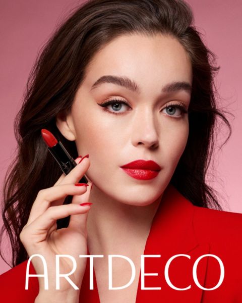 Eine Frau mit heller Haut und dunklem Haar posiert vor einem rosa Hintergrund, hält einen roten Lippenstift in der Hand und trägt einen roten Blazer sowie einen dazu passenden roten Lippenstift. Darunter steht der Schriftzug „ARTDECO“.