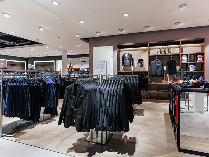 Ein modernes Bekleidungsgeschäft mit Kleiderständern voller ordentlich aufgehängter Anzüge. Auf den Regalen im Hintergrund sind gefaltete Hemden, Blazer und Accessoires zu sehen. Das Geschäft ist hell beleuchtet und hat ein elegantes, übersichtliches Layout.