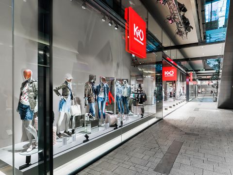 Ein Schaufenster eines Modegeschäfts, in dem Schaufensterpuppen in trendigen Freizeitoutfits, darunter Jacken und Jeansshorts, ausgestellt sind. Das Geschäft hat große Glasfenster und befindet sich in einem modernen Einkaufszentrum mit eleganter Architektur.