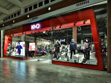 Schaufenster von K&Ö in einem Einkaufszentrum mit Schaufensterpuppen in verschiedenen Kleidungsstilen. Die Ausstellung umfasst Jacken, Hemden und Accessoires unter heller Beleuchtung, im Inneren sind weitere Kleidungsstücke zu sehen.