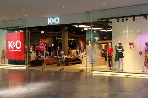 Schaufenster von Kastner & Öhler mit großen Glasfenstern, in denen bekleidete Schaufensterpuppen zu sehen sind. Im Ladeninneren sind Kleiderständer und eine Auswahl an Bekleidung zu sehen. Das K&O-Logo leuchtet deutlich sichtbar über dem Eingang.