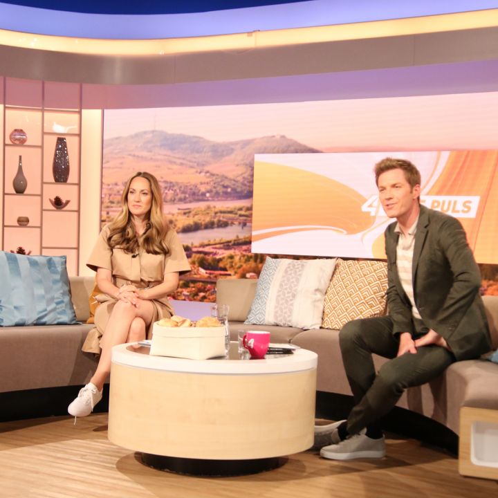 Ein Mann und eine Frau sitzen an einem modernen Fernseher mit einem U-förmigen Sofa und dekorativen Kissen. Auf einem Couchtisch stehen eine Schachtel und eine Tasse. Im Hintergrund ist ein malerisches Landschaftsbild zu sehen. Von oben sind Studiolichter zu sehen.