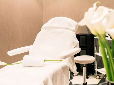 Ein Spa-Behandlungsraum mit einem weiß bezogenen Liegestuhl, einem aufgerollten Handtuch und einer langstieligen Blume. Im Hintergrund stehen ein Hocker und ein Regal. Im Vordergrund sind weiße Blumen zu sehen.