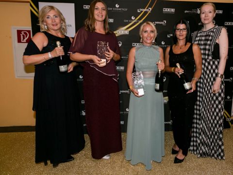 Eine Gruppe von fünf Frauen in Abendgarderobe steht lächelnd zusammen und hält Auszeichnungen und Flaschen vor einem Hintergrund mit verschiedenen Logos.