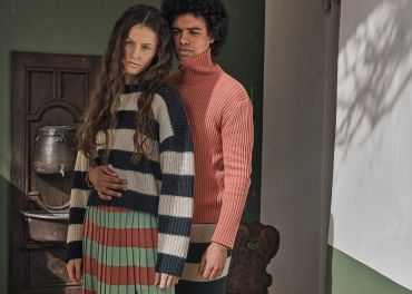 Eine Frau mit langen Haaren trägt einen gestreiften Pullover und Rock und steht vor einem Mann in einem rosa Rollkragenpullover. Sie posieren drinnen neben einer rustikalen Holztür, wobei sanftes Tageslicht Schatten an die Wand wirft.