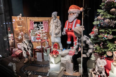 In einem festlichen Schaufenster sind Schaufensterpuppen in Winterkleidung zu sehen, umgeben von Geschenkkartons, Spielzeug und einer großen Weihnachtsmannfigur. Auf der rechten Seite steht ein geschmückter Weihnachtsbaum, der das Weihnachtsthema noch verstärkt.