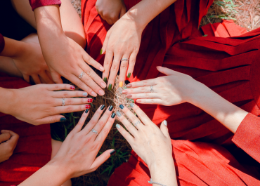 Eine Gruppe von Menschen sitzt im Kreis und streckt ihre Hände nach innen aus. Jede Person trägt ein rotes Kleid und ihre Hände sind mit Ringen und lackierten Nägeln geschmückt. Der Hintergrund ist grasbewachsen.