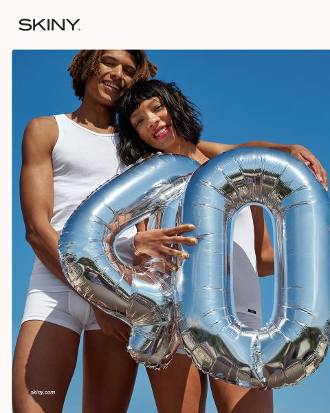 Zwei Personen in weißer Unterwäsche halten lächelnd einen silbernen Luftballon mit der Zahl "40" vor einem strahlend blauen Himmel.