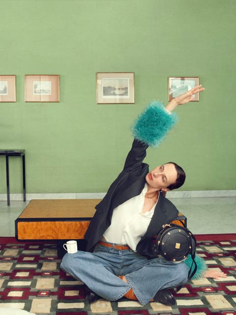 Eine Person in Jeans und einem Blazer mit blauen, pelzigen Manschetten posiert mit erhobenem Arm in einem stilvollen Raum mit grünen Wänden, gerahmten Kunstwerken und einem gemusterten Teppich.
