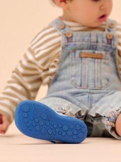 Ein Kleinkind in gestreiftem Hemd und Jeans-Overall sitzt mit einem Bein im Freien und zeigt die strukturierte blaue Sohle seines Schuhs.