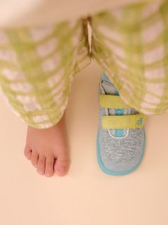 Ein nackter Kinderfuß neben einem Turnschuh mit gelben Riemen, beide sichtbar unter einer hellgrün karierten Hose auf einem beigen Boden.