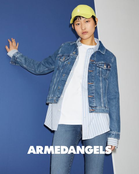 Person posiert vor einem blau-weißen Hintergrund, trägt eine Jeansjacke über einem gestreiften Hemd, blaue Jeans und eine limettengrüne Mütze. Unten steht in weißer Schrift das Wort „ARMEDANGELS“.