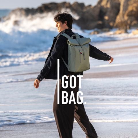 Eine Person geht an einem Strand mit Wellen im Hintergrund entlang und trägt einen hellgrünen Rucksack. Der Text lautet "I GOT BAG.