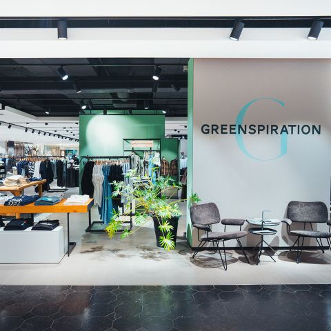 Modernes Bekleidungsgeschäft mit dem Schriftzug "GREENSPIRATION" an der Wand, stilvollen Sitzgelegenheiten, Grünpflanzen und ordentlich ausgestellten Kleiderständern.