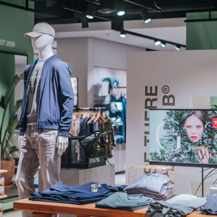 Auslage eines Bekleidungsgeschäfts mit einer Schaufensterpuppe in Freizeitkleidung, gefalteter Kleidung auf einem Tisch, Schuhen in Regalen und einem Werbefoto im Hintergrund.