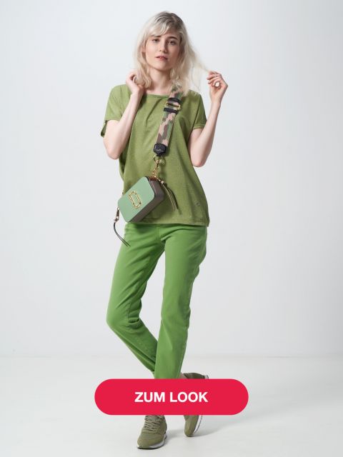 Eine Person mit hellem Haar trägt ein grünes T-Shirt, grüne Hosen und grüne Turnschuhe und trägt eine kleine grüne Tasche mit einem Camouflage-Gurt. Der Hintergrund ist schlicht und unten ist ein rotes Banner mit dem Text „ZUM LOOK.