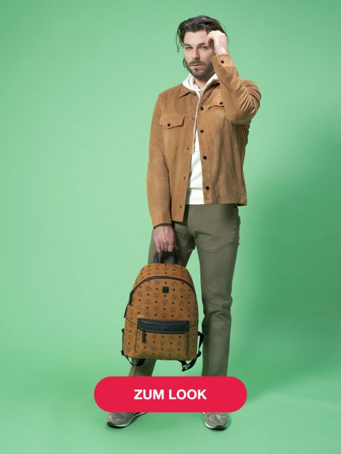 Ein Mann mit braunen Haaren steht vor einem grünen Hintergrund. Er trägt eine hellbraune Jacke, ein weißes Hemd und olivfarbene Hosen und hält einen braunen Rucksack mit schwarzem Muster in der Hand. Turnschuhe vervollständigen sein Outfit. Auf einem roten Button an der Unterseite steht „ZUM LOOK“.