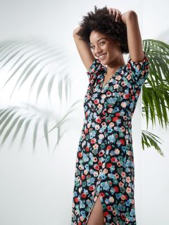 Eine Person lächelt und richtet ihre Haare. Sie trägt ein geblümtes Kleid mit Schlitz. Sie steht vor einem hellen Hintergrund, auf dem Palmblätter Schatten werfen.