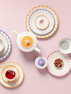 Bunte Teller mit Blumenmuster, eine Teekanne, eine Tasse mit Tee und Zitrone sowie ein Gebäckstück, arrangiert auf einem rosa Hintergrund.