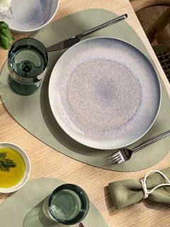 Tischgedeck mit einer grünen Tischunterlage, einem Keramikteller, einem Glas, einer Gabel, einem Messer, einer kleinen Schale mit Öl und Dill sowie einer grünen Stoffserviette mit Serviettenring.