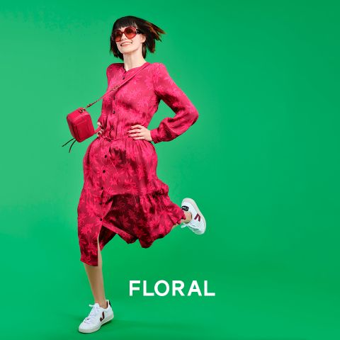 Eine Frau in einem fließenden rosa Blumenkleid und weißen Turnschuhen springt freudig vor einem leuchtend grünen Hintergrund. Sie trägt eine rote Sonnenbrille und hält eine kleine rosa Tasche in der Hand. Unten ist das Wort „FLORAL“ zu sehen.