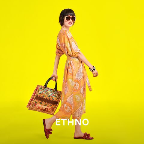 Eine Person mit Sonnenbrille und gelb gemustertem Kleid posiert selbstbewusst vor einem leuchtend gelben Hintergrund. Sie hält eine bunt gemusterte Tasche in der Hand und trägt rote Sandalen. Unten ist das Wort „ETHNO“ zu sehen.