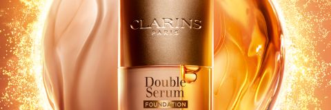 Eine Clarins Double Serum Foundation Flasche steht in der Mitte vor einem leuchtenden, goldenen, abstrakten Hintergrund mit einem Text, der die Foundation als revolutionär anpreist.