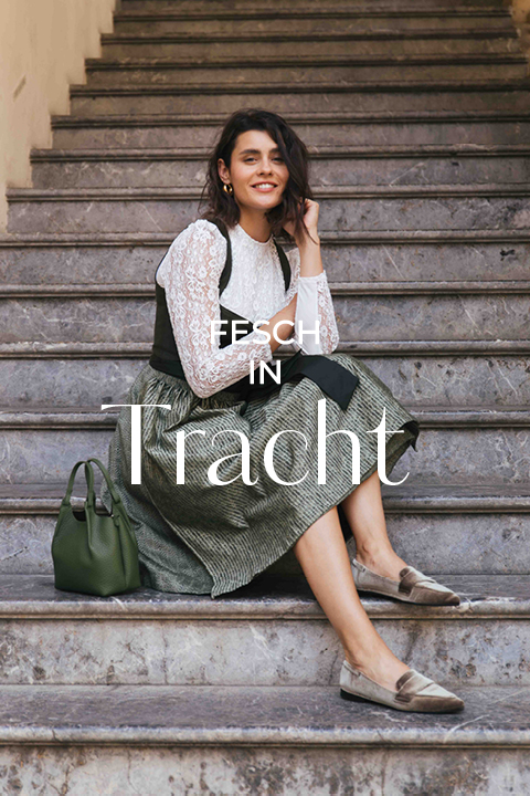 Eine Frau in einem traditionellen Kleid sitzt lächelnd auf einer Steintreppe, neben sich eine grüne Handtasche. Der Text lautet "Fesch in Tracht.