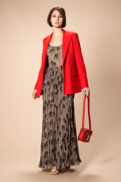Vor einem beigen Hintergrund steht eine Person, die ein gemustertes Maxikleid und einen leuchtend roten Blazer trägt und eine rote Handtasche hält. Sie hat kurze, dunkle Haare und blickt zur Seite.