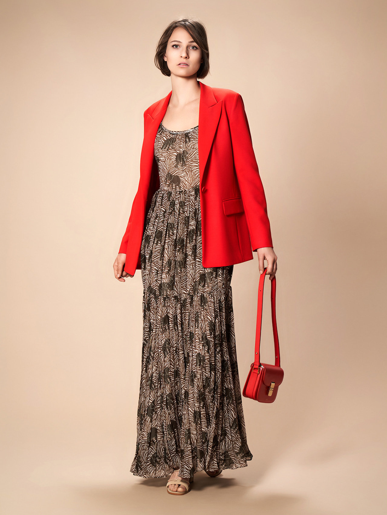 Vor einem beigen Hintergrund steht eine Person, die ein gemustertes Maxikleid und einen leuchtend roten Blazer trägt und eine rote Handtasche hält. Sie hat kurze, dunkle Haare und blickt zur Seite.