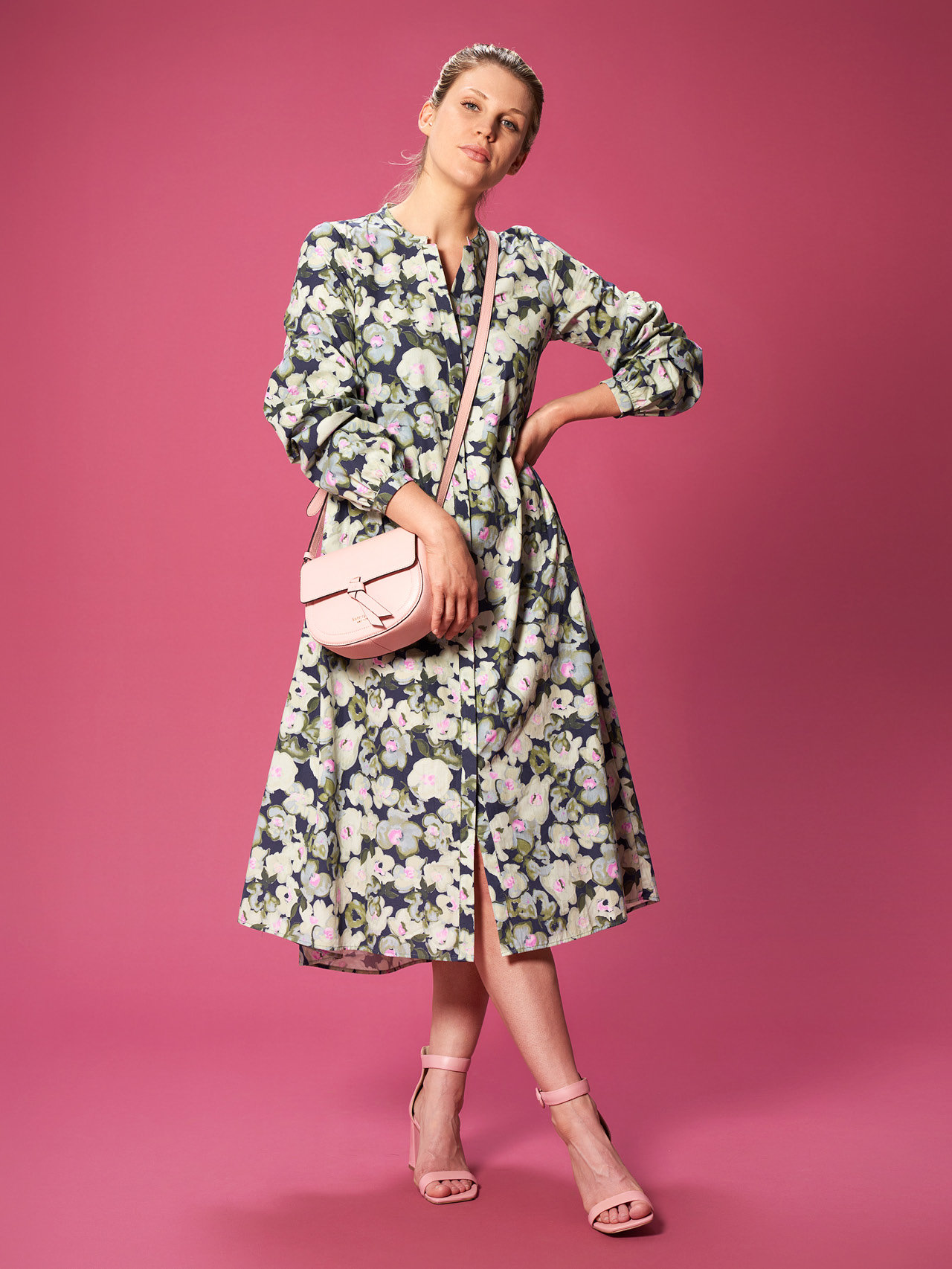 Eine Person steht vor einem rosa Hintergrund und trägt ein langärmeliges Kleid mit Blumenmuster. Sie hält eine hellrosa Tasche mit Riemen über der Schulter. Das Outfit wird durch hellrosa Sandalen mit hohen Absätzen komplettiert.