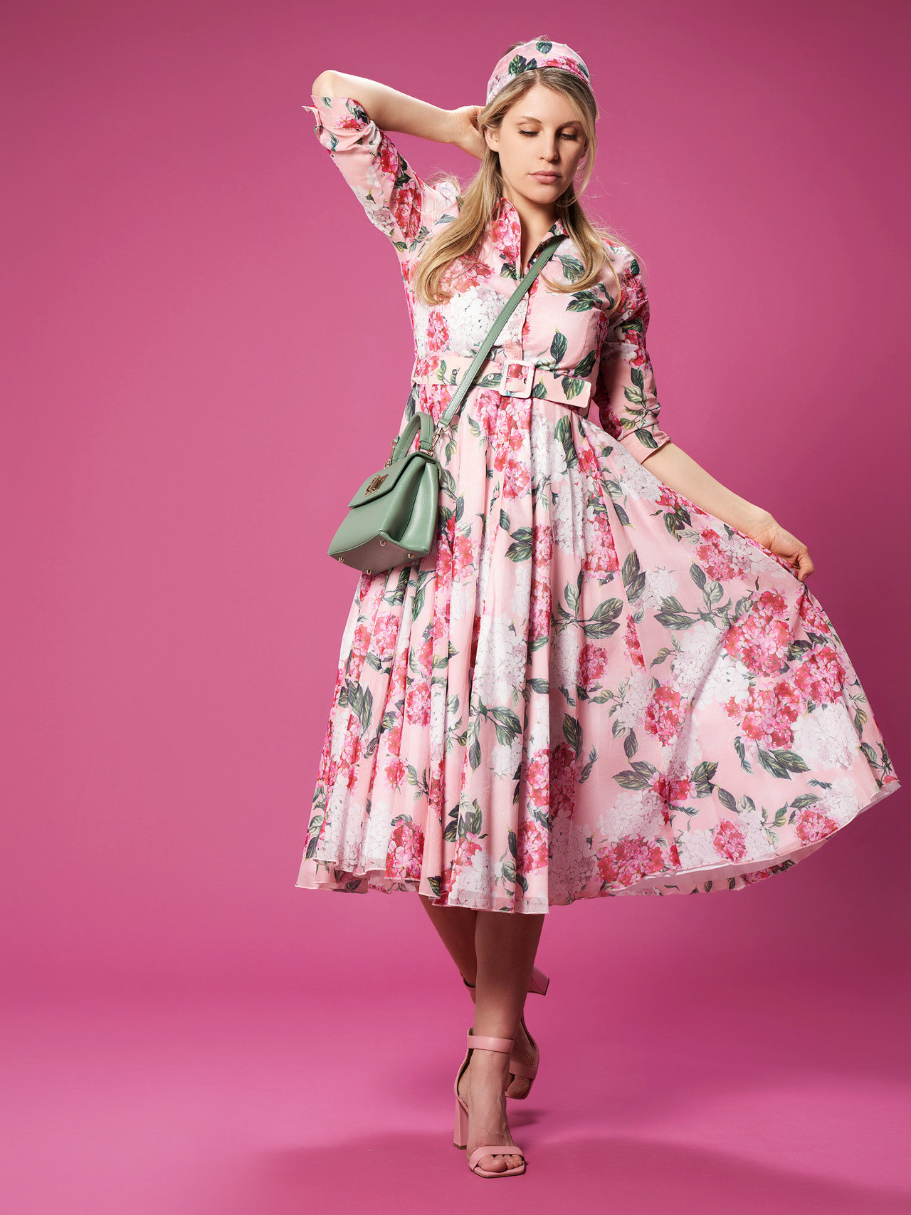Eine Frau posiert vor einem rosa Hintergrund und trägt ein rosa Kleid mit Blumenmuster und dazu passendem Haarband. Mit einer Hand hält sie den Rock hoch und mit der anderen berührt sie ihr Haar. Als Accessoires trägt sie eine grüne Umhängetasche und rosa High Heels.