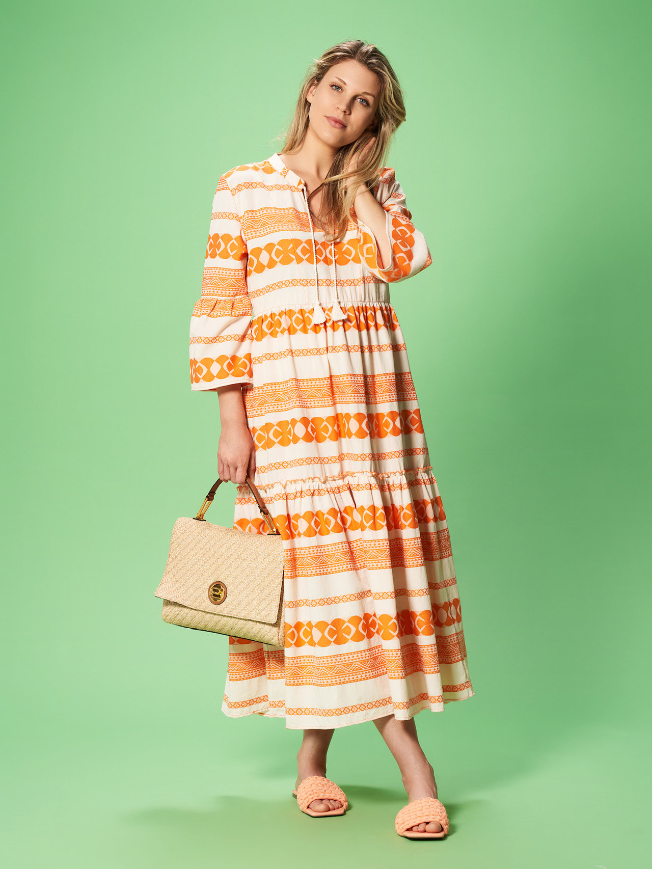 Eine Frau in einem langen, gemusterten Kleid mit orangen und weißen Mustern steht auf grünem Hintergrund. Sie hält eine beige gewebte Handtasche in einer Hand und trägt dazu passende Sandalen. Ihre Haare sind offen und sie blickt leicht zur Seite.