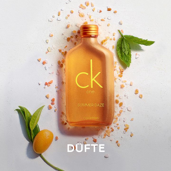 Eine orangefarbene CK One Summer Daze-Flasche, umgeben von verstreutem grobem Salz, einer kleinen Zitrusfrucht, grünen Blättern und einem kleinen Zweig auf weißem Hintergrund. Unten steht das Wort „DUFTE“.